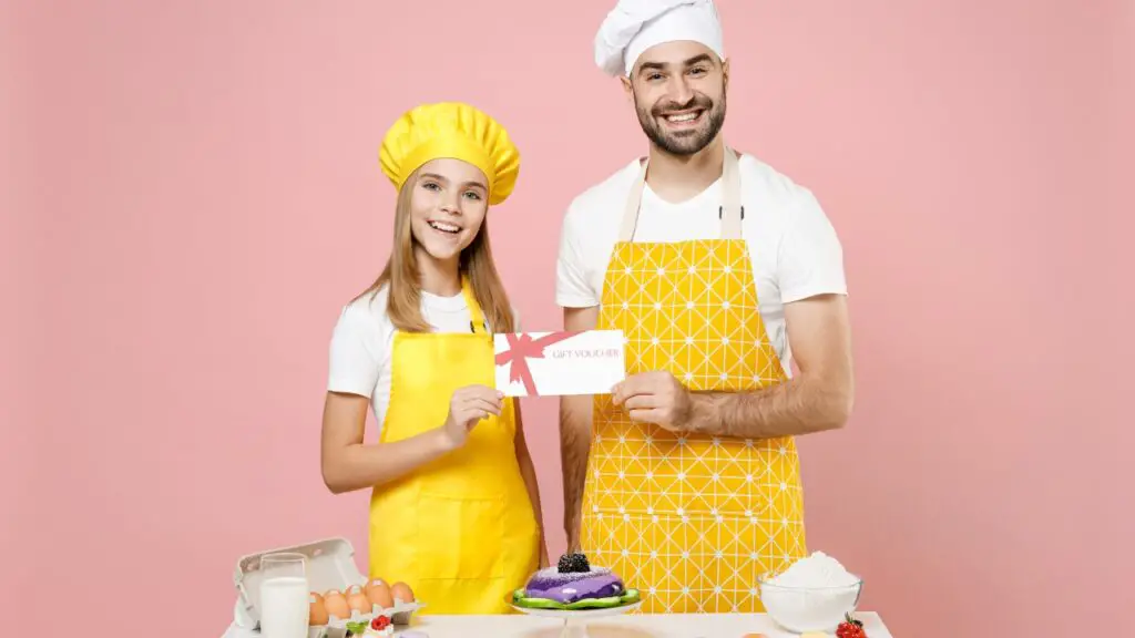 Man and girl in yellow aprons holding a gift voucher in a kitchen setup